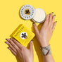 Two hands with black-tipped nails rest on a yellow surface next to an open tin and box of NOPALERA Moisturizing Botanical Bar. The person wears a beaded bracelet, and the plastic-free packaging shows bold black text and a woman’s face design.