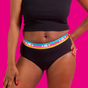 A person stands against a bright pink background wearing Here We Flos Flo Reusable Cotton Period Underwear—black, high-waisted, ultra-absorbent with a colorful waistband featuring hearts and HC letters—paired with a black crop top.