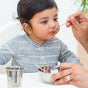 A baby in a gray striped shirt sits in a high chair as an adult feeds them orange baby food using Ahimsa Baby Starter Spoons from a small metal bowl.
