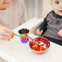 An adult hands an Ahimsa Baby Starter Spoon to a baby in a high chair, with a rainbow-colored bowl of chopped strawberries and a matching cup on the tray.