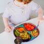 A young child enjoys a meal of chickpeas, carrot sticks, watermelon, pasta, and edamame served on the Ahimsa Balanced Bites Divided Plate—a colorful stainless steel option perfect for balanced meals.