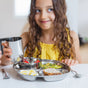 A young girl with long curly hair, wearing a yellow top, sits at a table smiling and holding an Ahimsa Mindful Mealtime Set stainless steel cup. In front of her is the matching tray with eggs, bread, vegetables, and strawberries.