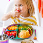 A young child in a yellow-striped shirt enjoys a berry from the colorful Ahimsa Mindful Mealtime Set, which is filled with fruit, cheese cubes, cucumber slices, and a sandwich.