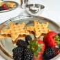 The Ahimsa Purposeful Plate holds two star-shaped waffles, fresh strawberries, and blackberries. A fork, another plate with waffles and berries, and a small metal bowl appear in the background on a white surface.