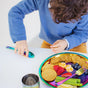 A child in a blue sweater sits at a table with the Ahimsa Purposeful Plate, featuring crackers, cheese, strawberries, blueberries, celery, and a cup. The child reaches for a colorful utensil while looking down.