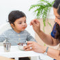 A woman feeds a young child in a gray shirt seated in a high chair. On the tray are Ahimsa Simple Serve Bowls Set and a metal cup. A green plant is visible in the background.