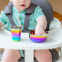 A baby in a green knitted outfit and safety harness uses the Ahimsa Simple Serve Bowls Set—colorful, rainbow-metal stainless steel bowls with a matching cup. Both are dishwasher-safe for easy cleaning.