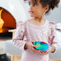 A young child in a pink dress with gold patterns holds the Ahimsa Smart Snacking Bowl, standing indoors near a lit fireplace and a gray sofa.