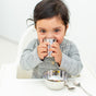 A young child with dark hair, in a gray shirt, sits in a high chair sipping from an Ahimsa Starter Sips Cup. In front of them on the table are a metal bowl with food and a spoon.