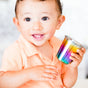 A smiling toddler in a peach shirt holds a shiny, rainbow-colored metal cup, looking up with bright eyes—showcasing Ahimsa’s Starter Sips Cups, non-toxic and designed for safe, everyday use.