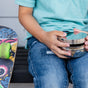 A child in a light blue shirt and jeans sits on a bench, holding Ahimsa’s The Large Circle - Stainless Steel Food Storage Container. A colorful skateboard with green wheels is beside them.