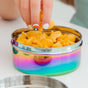 A hand with rainbow nail art picks up a goldfish cracker from Ahimsa's The Large Circle, a stainless steel food storage container filled with similar crackers.