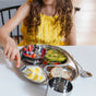 A child in a yellow dress sits at a table with food in a metal plate, holding a fork from the Ahimsa Utensil Set, ready to eat. A matching spoon and cup are nearby.