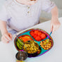 A young child sits at a table, ready to eat with the Ahimsa Utensil Set, using a spoon to enjoy food from a colorful divided plate filled with watermelon, chickpeas, crackers, and edamame.