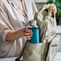 A person puts a Black + Blum Glass Water Bottle with a silicone sleeve into the side pocket of a light beige backpack, wearing a light shirt with indoor plants in the background.
