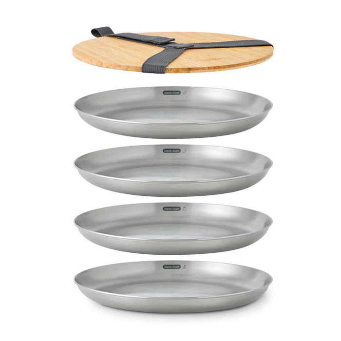 The Black + Blum Steel Plate Set of 4 is stacked neatly beneath a bamboo chopping board with a black carry strap, all arranged vertically on a white background.