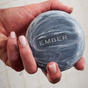 A hand holds a round, dark gray Charcoal Exfoliating Cleanser Bar from Ember Wellness, with water droplets on its surface; the word "EMBER" is engraved on the bar and a tiled wall forms the background.