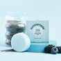 A pastel blue background displays the Sunniemade Smoothing Conditioner Bar - Chia Glow, formulated with nourishing chia seed oil to help reduce frizz, positioned next to its box alongside chia seeds, a glass jar, blackberries, and a blue circular prop.