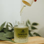 A dropper holds oil above an open bottle labeled Dewdrop Cleansing Oil by Hearth and Homestead, a nourishing formula designed to effortlessly dissolve makeup as an effective remover. Green leaves and a wooden surface are visible in the background.