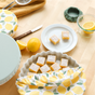 A table displays lemon bars with powdered sugar, fresh lemons, a knife, a glass jar, and sustainable food storage using Esembly Baby's Reusable Bowl Covers made from Upcycled Materials (4-Pack).