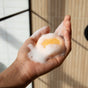 A hand holding a small Sunniemade Moisturizing Shampoo Bar - Mighty Mango, covered in white foam, with a blurred light brown and white tiled background.