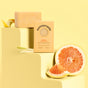 A bar of Sunniemade’s Moisturizing Hand & Body Bar Soap—Main Squeeze, its packaging, grapefruit halves, lemon peel, and lemongrass are arranged on yellow blocks against a yellow background—an inviting display that celebrates natural body care.