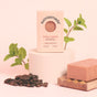 A bar of Sunniemade Exfoliating Body Bar Soap - Rise & Grind is shown with coffee beans, a wooden soap holder, and fresh mint sprigs against a soft pink backdrop. Infused with coffee grounds for gentle exfoliation and natural body care, the upright soap packaging is displayed on a round pedestal.