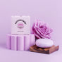 A vibrant purple Strengthening Shampoo Bar – Super Bloom by Sunniemade, enriched with rosehip oil, sits atop a wooden tray beside a pastel pink Sunniemade Super Bloom box and an elegant lavender rose—all artfully displayed on lilac platforms with a soft pastel purple backdrop.
