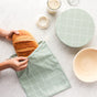 Hands place a freshly baked loaf of sourdough into a green, checkered bag from the Sourdough Proofing Kit by Esembly Baby, set on a white surface. Nearby are items from the kit, including a jar, round cloth-covered container, and woven bowl.