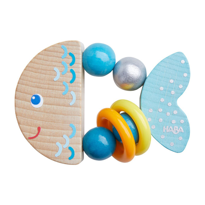 The HABA Rattlefish Wooden Baby Rattle is shaped like a smiling fish with blue scales, yellow rings, blue beads, and a dotted tail fin. HABA is on the tail; ideal as a sensory clutching toy for babies.
