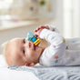 A baby in a white onesie with colorful stars lies on a soft surface, smiling and holding a HABA Rattlefish Wooden Baby Rattle—an engaging blue and yellow sensory toy.
