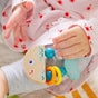 A baby in a pink outfit with red flower patterns holds the HABA Rattlefish Wooden Baby Rattle—a colorful sensory toy featuring blue scales, orange rings, and blue beads.