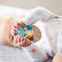 A baby in a white onesie with pastel stars holds the colorful HABA Rattlefish Wooden Baby Rattle while lying on a soft, light gray surface. The babys face is partially visible, highlighting the vibrant rattle in their hands.