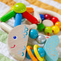 The HABA Rattlefish Wooden Baby Rattle, featuring bright colors and smiling faces, lies on a striped quilted surface with colorful beads in blue, yellow, and green.