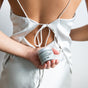 A person in a white satin dress with an open back holds a jar of Little Seed Farm Deodorant Creams behind their back, highlighting the product and the elegant ties of the dress.