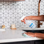 A person uses Meliora All-Purpose Home Cleaner Refill Tablets, spraying the cleaner onto a white kitchen countertop and wiping with a cloth. In the background are geometric white tiles and a copper faucet.