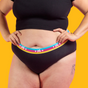 Someone poses against a solid orange background wearing Flo Reusable Cotton Period Underwear by Here We Flo, featuring a colorful waistband with hearts and “HC”, paired with a black crop top. A small upper thigh tattoo is visible.