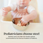 A baby sits in a high chair reaching into an Ahimsa Simple Serve Bowl filled with snacks. Text below: Pediatricians choose steel. Stainless steel is the ONLY kid-friendly material recommended by the American Academy of Pediatrics.