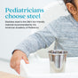 A child in a gray sweater sits at a white table, reaching for an Ahimsa Starter Sips Cup. Text reads: Pediatricians recommend stainless steel—it's the only kid-friendly material endorsed by the American Academy of Pediatrics.