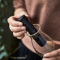 A person places a Black + Blum Active Charcoal Water Filter into a clear glass carafe, holding the stick with one hand and the carafe with the other, against a softly blurred background.