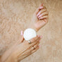 A person with long, light pink manicured nails holds a round NOPALERA Moisturizing Botanical Bar in one hand and touches their wrist with the other, set against a textured beige background.