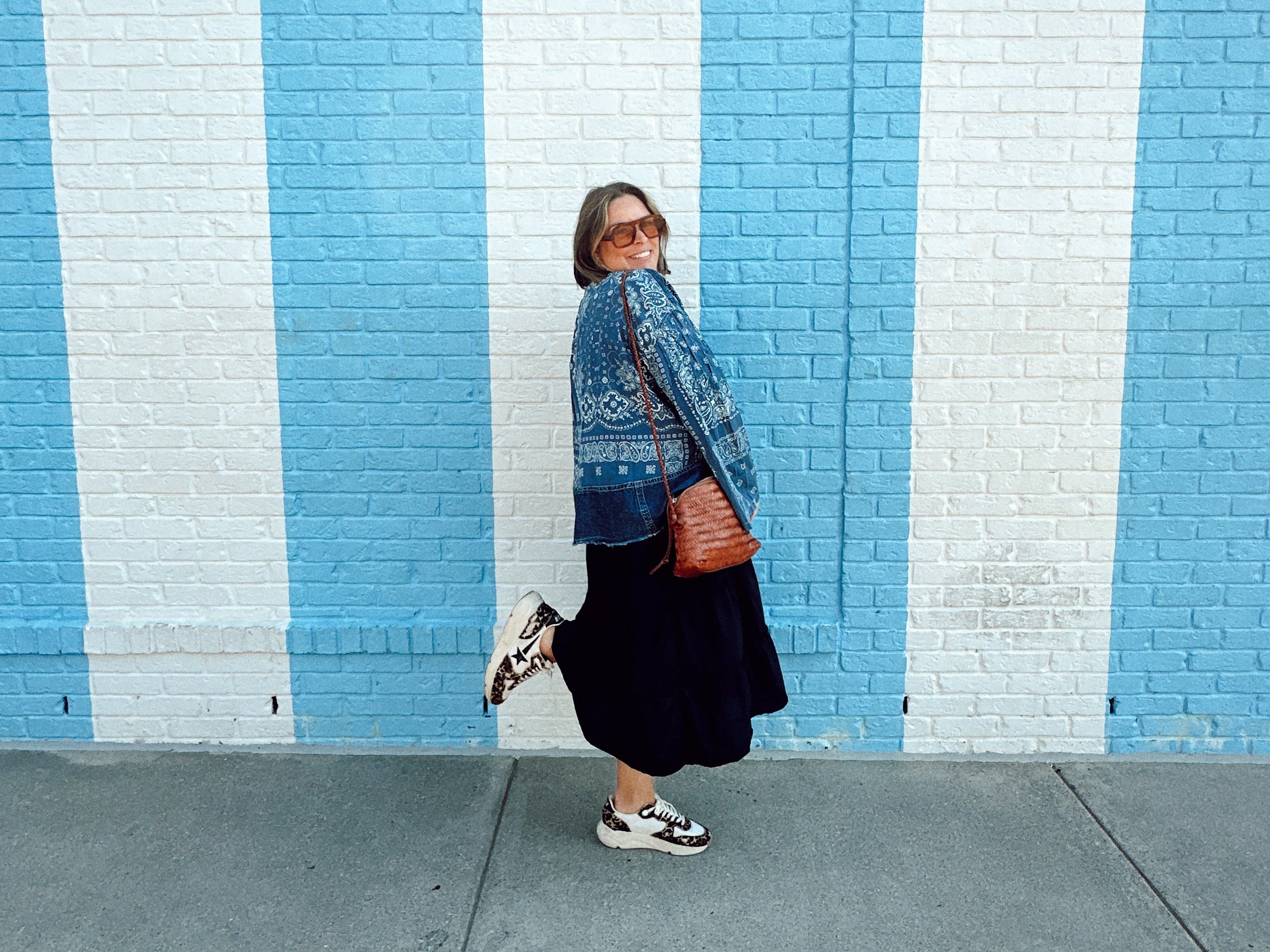 A person wearing sunglasses, a patterned blue sweater, black skirt, and white sneakers poses playfully against a brick wall painted with vertical blue and white stripes. They have one foot raised and carry a brown bag.