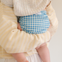 A person in a light yellow striped sweater holds a baby wearing Esembly Baby's Outer Diaper Cover + Swim Diaper (Size 1, 100% OEKO-TEX certified recycled fabric), with the baby's face gently covered by a knitted blanket.