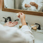 A person in a white robe dispenses Hearth and Homestead’s Dewdrop Cleansing Oil from a dropper onto their palm over a bathroom sink, with toiletries and a mirror visible in the background.