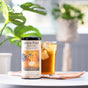 A tin of The Republic of Tea's Ginger Peach Black Tea, known for its natural flavors and digestive benefits, sits before a lemon-iced tea glass, surrounded by lush green plants in gentle sunlight.