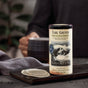 A cylindrical tin of Earl Greyer Black Tea by The Republic of Tea sits on a tray with tea bags, as a person holds a mug in the blurred background. The label features an elephant and notes its citrus flavor from natural bergamot oil.