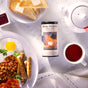 A breakfast scene featuring The Republic of Tea’s British Breakfast Black Tea tin, a teapot, cup of tea, buttered toast, jam, beans, sausages, fried eggs, and sliced tomatoes on a white marble surface.
