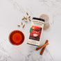 A tin of The Republic of Tea Cardamon Cinnamon Herbal Tea, tea bags, a glass cup of tea, cinnamon sticks, and cardamom pods—classic Indian spices—are beautifully arranged on a white marble surface.