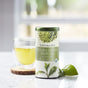 A tin of The Republic of Tea Organic Double Green Matcha Tea USDA rests on marble beside a glass cup of Japanese tea on a wooden coaster, with tea leaves nearby and a bright, blurred background.