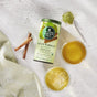 A canister of The Republic of Tea Organic Lean Green SuperGreen Tea rests on a white surface, surrounded by two cups of green tea, cinnamon sticks, a spoonful of matcha powder, and a light green patterned cloth.